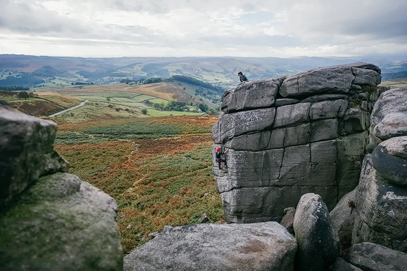 Twee mensen die aan het rotsklimmen zijn op een grote rotsformatie in een schilderachtig landschap met glooiende heuvels, groene velden en een bewolkte hemel op de achtergrond.